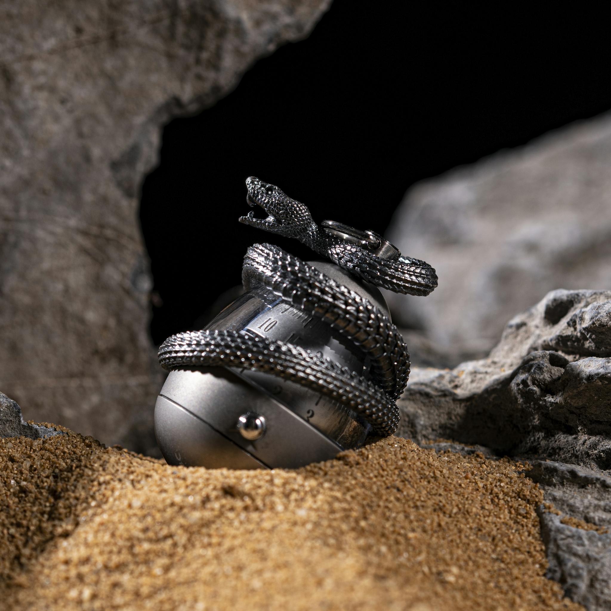 Artistic display of silver snake jewelry amidst sand and rocks, highlighting intricate details.